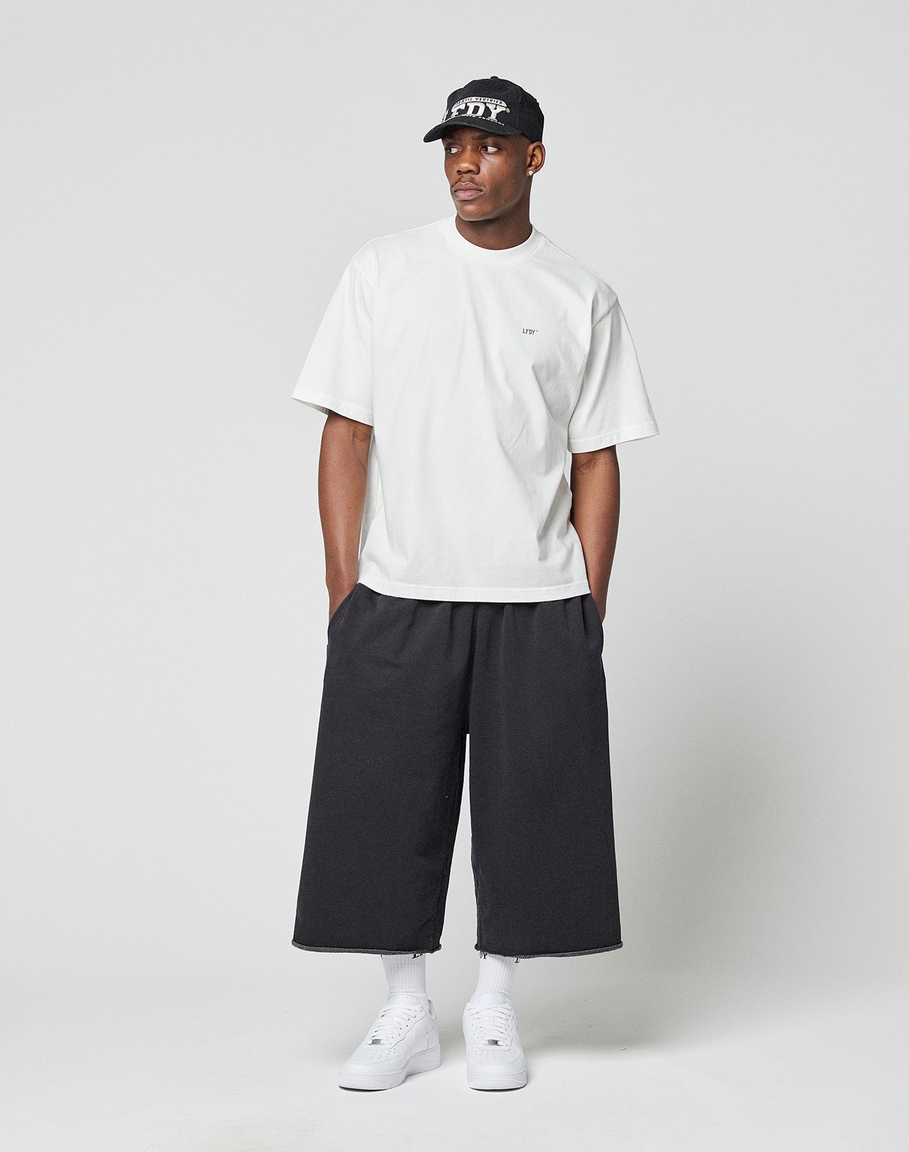 A man stands against a plain background wearing the LIVE FAST DIE YOUNG LFDY Zizou Tee, black wide-leg cropped pants, white socks and sneakers, and a black baseball cap. Hands in his pockets, he looks to the side.