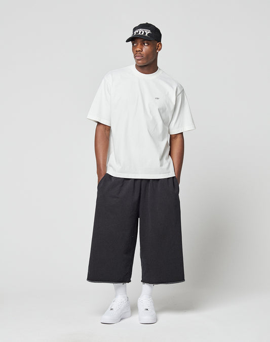 A man stands against a plain background wearing the LIVE FAST DIE YOUNG LFDY Zizou Tee, black wide-leg cropped pants, white socks and sneakers, and a black baseball cap. Hands in his pockets, he looks to the side.