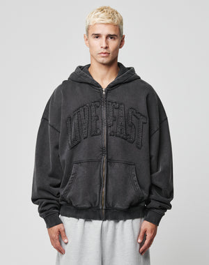 A young man with short blond hair wears the LIVE FAST DIE YOUNG Ripped College Hooded Zip, a loose black hoodie with textured LIVE FAST on the front, made from 100% Baumwolle, and light gray sweatpants against a plain gray background.