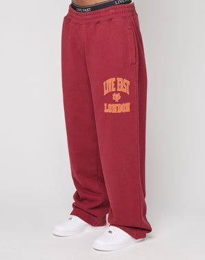 Lower body view of a person in LIVE FAST DIE YOUNG’s Live Fast London Wide Jogger—loose-fitting red sweatpants featuring “LIVE FAST LONDON” and a yellow lion graphic on the left leg—paired with white sneakers.