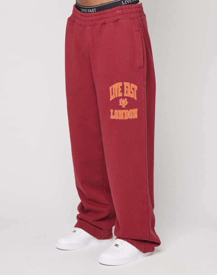 Lower body view of a person in LIVE FAST DIE YOUNG’s Live Fast London Wide Jogger—loose-fitting red sweatpants featuring “LIVE FAST LONDON” and a yellow lion graphic on the left leg—paired with white sneakers. | model