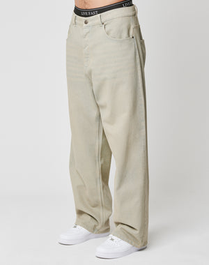 A person wears LFDY Baggy Denim by LIVE FAST DIE YOUNG—loose-fitting, light beige jeans with a visible black waistband featuring LIVE FAST text. Styled with white sneakers, made from hochwertige Baumwolle, against a plain background.