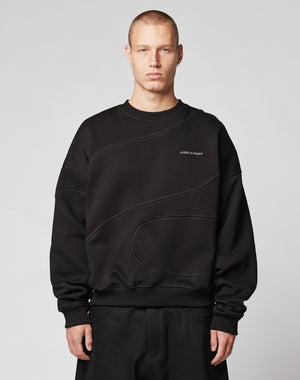 A person with short hair wears the LIVE FAST DIE YOUNG LFDY Wavy Sweater, an oversize black cotton sweatshirt with subtle curved stitching and a bold LIVE FAST print, standing against a plain light gray background.