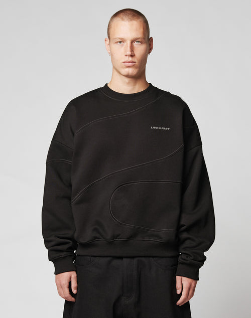 A person with short hair wears the LIVE FAST DIE YOUNG LFDY Wavy Sweater, an oversize black cotton sweatshirt with subtle curved stitching and a bold LIVE FAST print, standing against a plain light gray background.