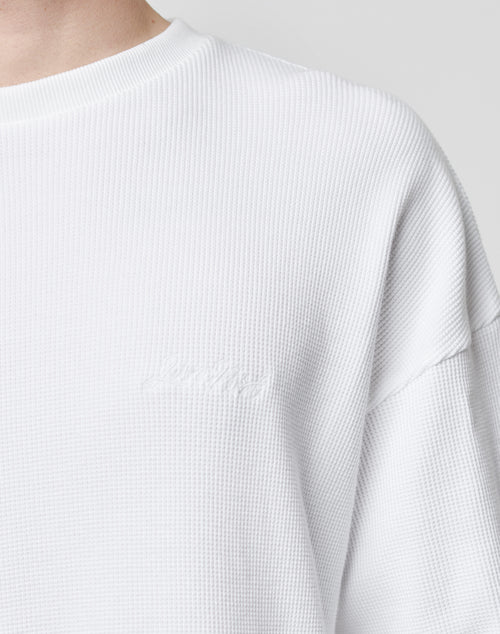 Close-up of a person wearing the LIVE FAST DIE YOUNG Basic Script Waffle Longsleeve, a textured white crew-neck shirt with an oversize fit and subtle logo embroidery near the chest. The background is plain and light-colored.