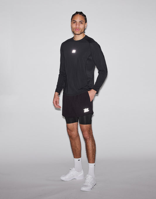 A man poses against a plain background, wearing the LIVE FAST DIE YOUNG Live Fast Performance Longsleeve in black with a reflective front print, black shorts over compression shorts, plus white socks and sneakers. He gazes neutrally at the camera.