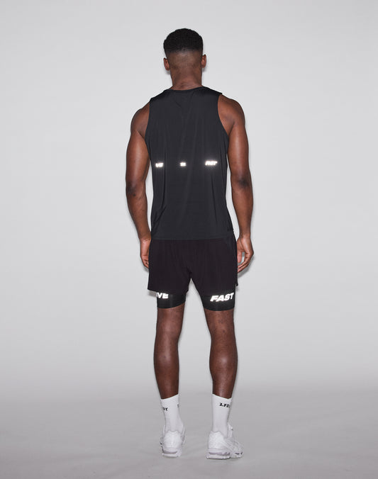 A man faces away from the camera, wearing a black LIVE FAST DIE YOUNG Live Fast Performance Tank, black shorts with FAST and LIVE in reflective print, white socks, and white sneakers against a plain light background.
