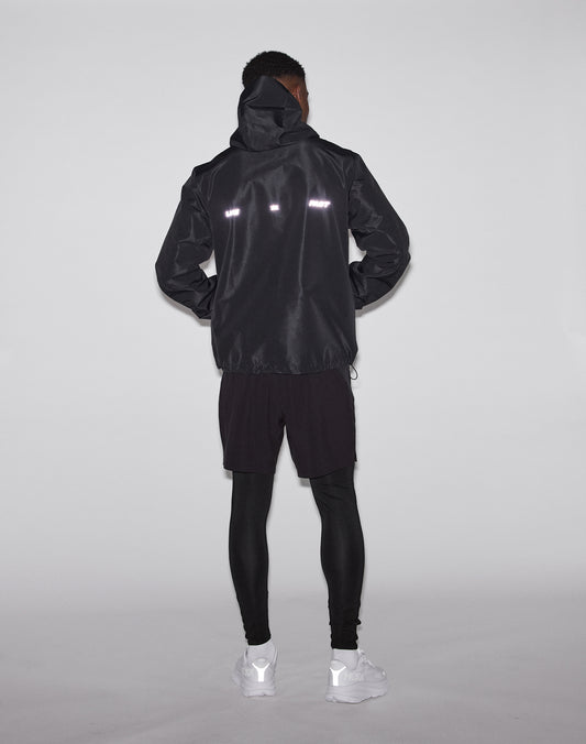 A person stands facing away, wearing the LIVE FAST DIE YOUNG Live Fast Performance Rain Jacket with waterproof seams and reflective strips, black shorts over leggings, and white sneakers against a plain light background.