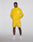 A man stands against a plain background wearing the bright yellow Live Fast Performance Running Jacket by LIVE FAST DIE YOUNG with reflektierender Frontprint, matching shorts, white socks, and white sneakers. Hands in pockets, he looks at the camera.
