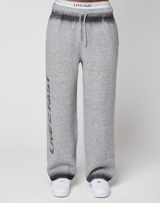 A person wears LIVE FAST DIE YOUNG’s Live Fast Boucle Pants in grey with a wide fit, straight leg, elastic waistband, and “LIVE FAST” printed vertically, paired with white sneakers, all shown against a plain background.