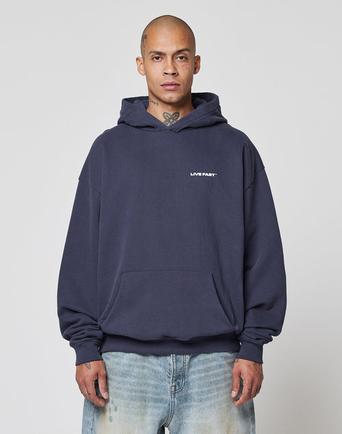 A person with a shaved head wears the Live Fast Plain Hooded by LIVE FAST DIE YOUNG in dark blue, featuring an oversize fit and front print, paired with light blue jeans, standing against a plain light background.