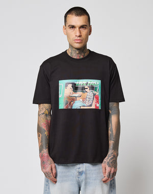 A man with neck and arm tattoos wears the LIVE FAST DIE YOUNG LFDY Sixers Tee, a regular fit black cotton T-shirt with a vibrant two-people-at-a-table graphic. He pairs it with light blue jeans against a white background. Care instructions on label.