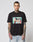 A man with neck and arm tattoos wears the LIVE FAST DIE YOUNG LFDY Sixers Tee, a regular fit black cotton T-shirt with a vibrant two-people-at-a-table graphic. He pairs it with light blue jeans against a white background. Care instructions on label.
