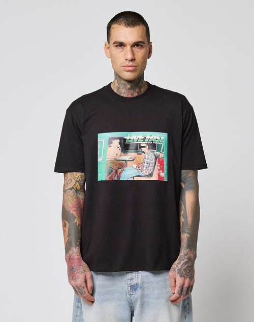 A man with neck and arm tattoos wears the LIVE FAST DIE YOUNG LFDY Sixers Tee, a regular fit black cotton T-shirt with a vibrant two-people-at-a-table graphic. He pairs it with light blue jeans against a white background. Care instructions on label.