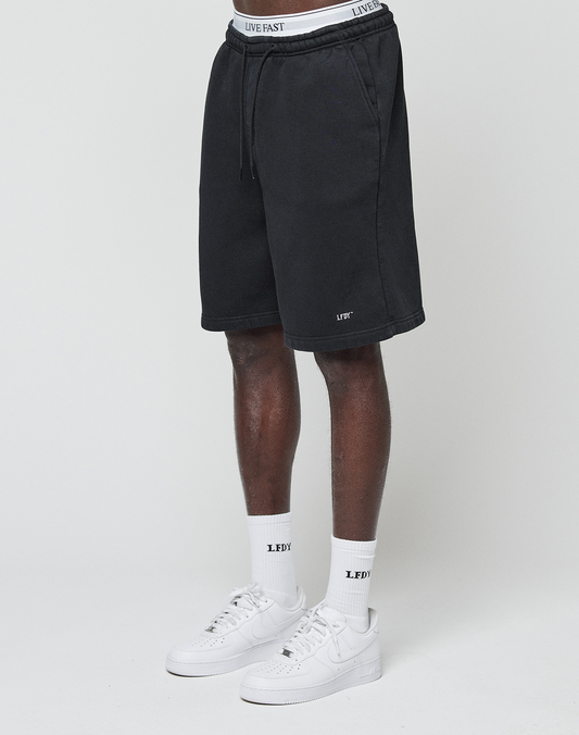 A person in LIVE FAST DIE YOUNG Basic Shorts with a logo print and elastic waistband, white crew LEDV socks, and white Nike sneakers stands against a white background. The waist-up photo highlights the shorts' distinctive branding.