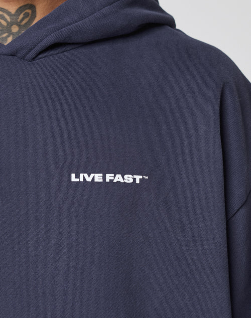 A person wears the Live Fast Plain Hooded by LIVE FAST DIE YOUNG, a dark navy Oversize Fit hoodie with a bold LIVE FAST™ front print; part of a neck tattoo is visible above the collar.