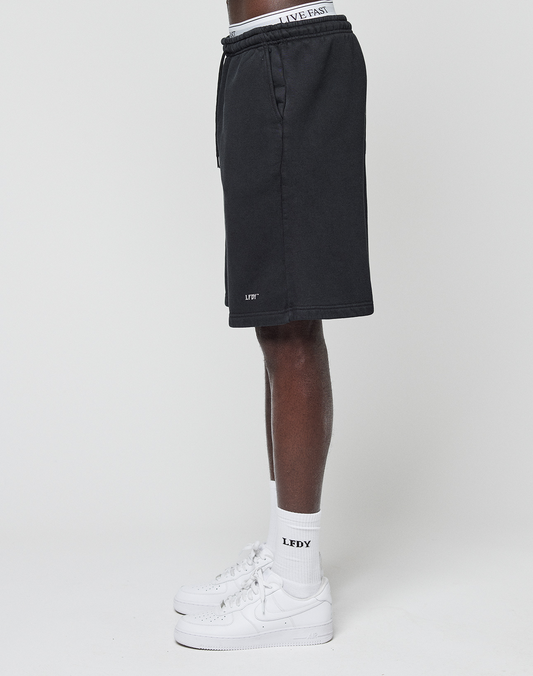 A person stands sideways against a plain white background wearing LIVE FAST DIE YOUNG Basic Shorts with a logo print near the hem and an elastic waistband that says LIVE FAST, paired with white sneakers and LFDY socks.