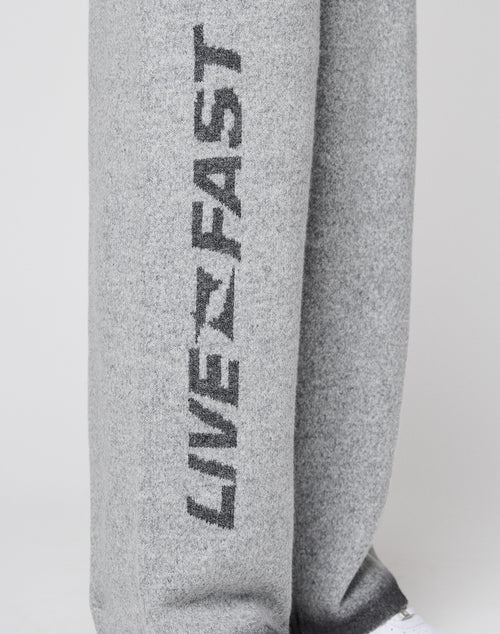 Close-up of LIVE FAST DIE YOUNG’s Live Fast Boucle Pants in light gray with straight leg and elastic waistband, featuring bold, vertical "LIVE FAST" print on one leg; partial white shoe visible at the bottom.