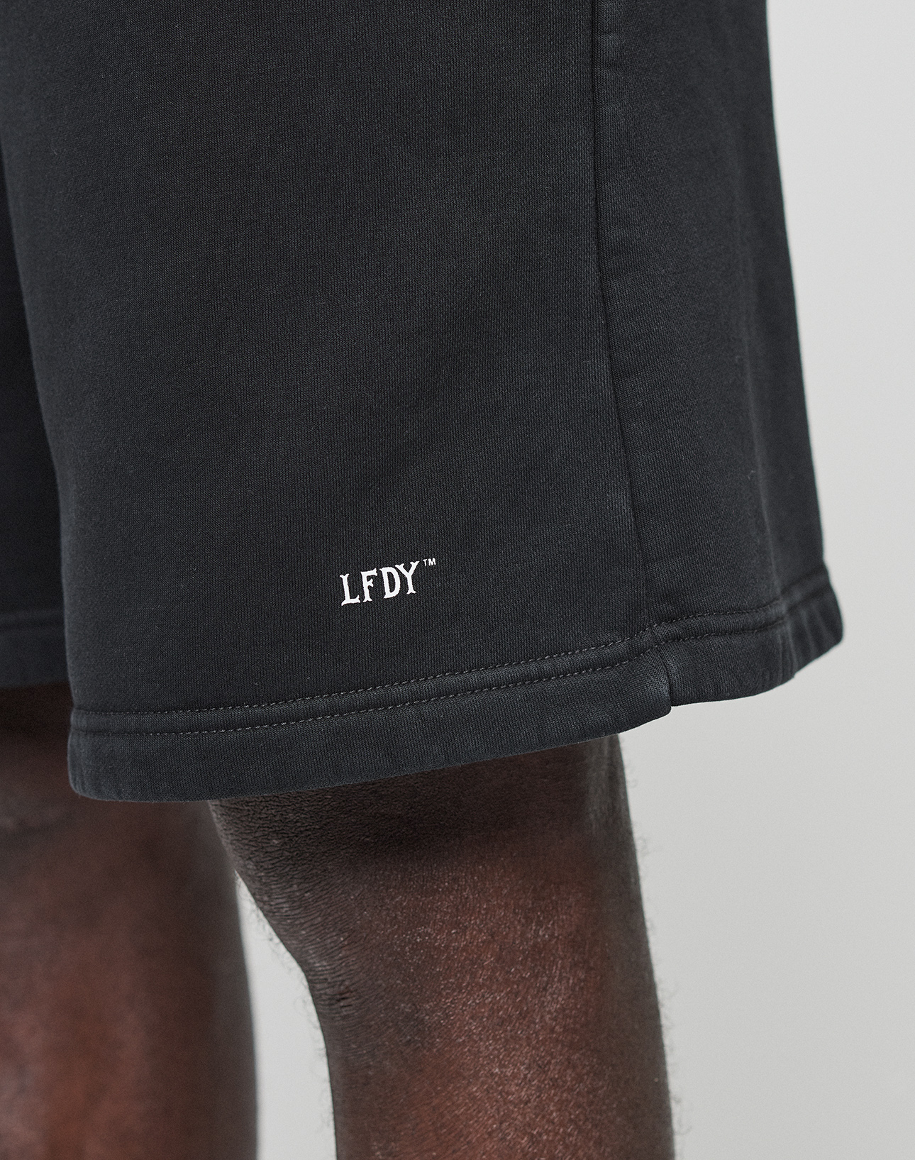 Close-up of a person wearing LIVE FAST DIE YOUNG Basic Shorts in black, featuring a regular fit and white LFDY™ logo embroidered near the hem. Only part of the shorts and the person’s upper leg are visible.