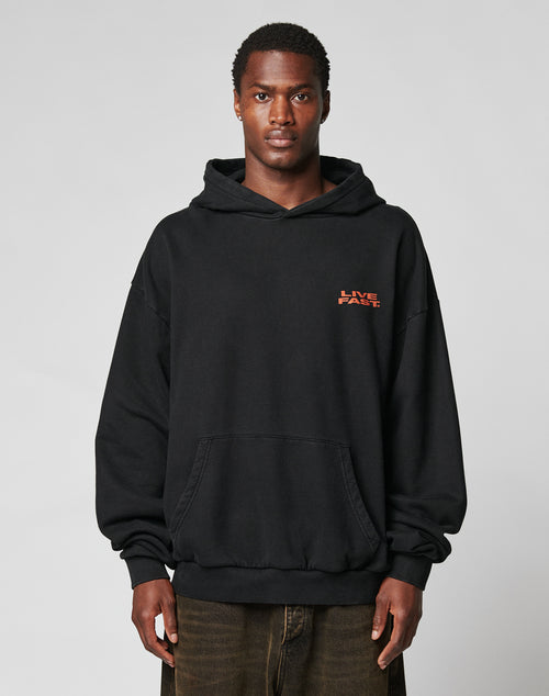 A man faces forward in the Print Over Hooded by LIVE FAST DIE YOUNG, an Oversize Fit black hoodie with a red LIVE FAST logo on the left chest, made from 100% cotton. The background is plain light gray.