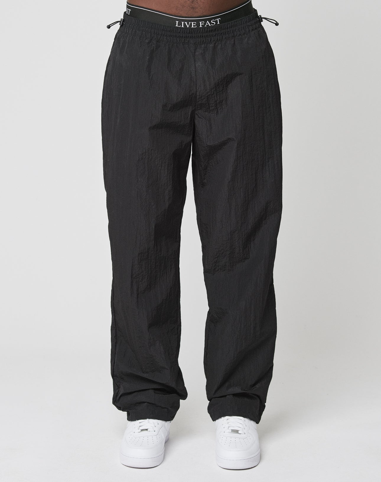 A person wears LIVE FAST DIE YOUNGs Live Fast Nylon Jogger—loose black track pants with subtle texture and an LIVE FAST elastic waistband—paired with white sneakers, set against a plain light backdrop.