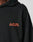 Close-up of a person in the Print Over Hooded by LIVE FAST DIE YOUNG, an oversize black hoodie made from 100% Baumwolle with LIVE FAST in bold orange on the left chest. Only part of the face and shoulder are visible against a neutral background.