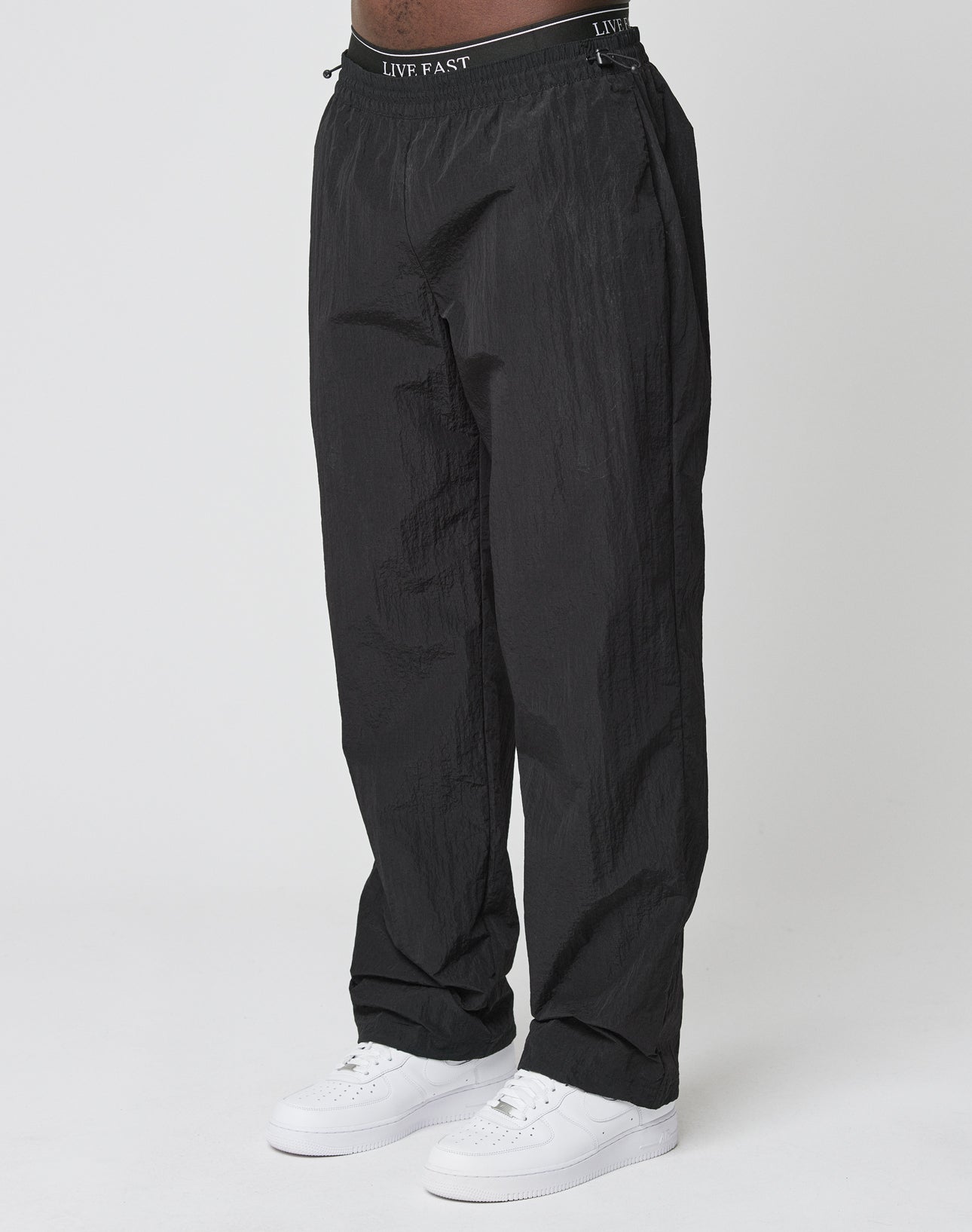 A person wears the black Live Fast Nylon Jogger by LIVE FAST DIE YOUNG, featuring an oversized fit, Elastikbund waistband with LIVE FAST text, paired with white sneakers, standing against a plain white background.