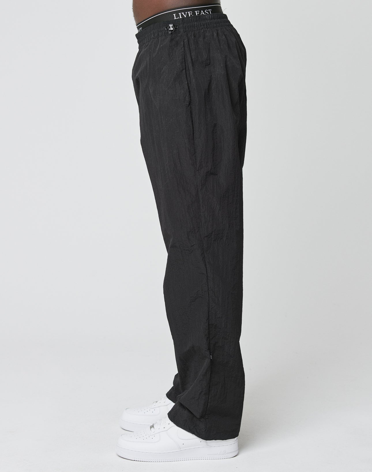 A person stands sideways against a plain white background wearing LIVE FAST DIE YOUNGs black, oversized Live Fast Nylon Jogger with an elastic waistband featuring LIVE FAST, paired with white sneakers.