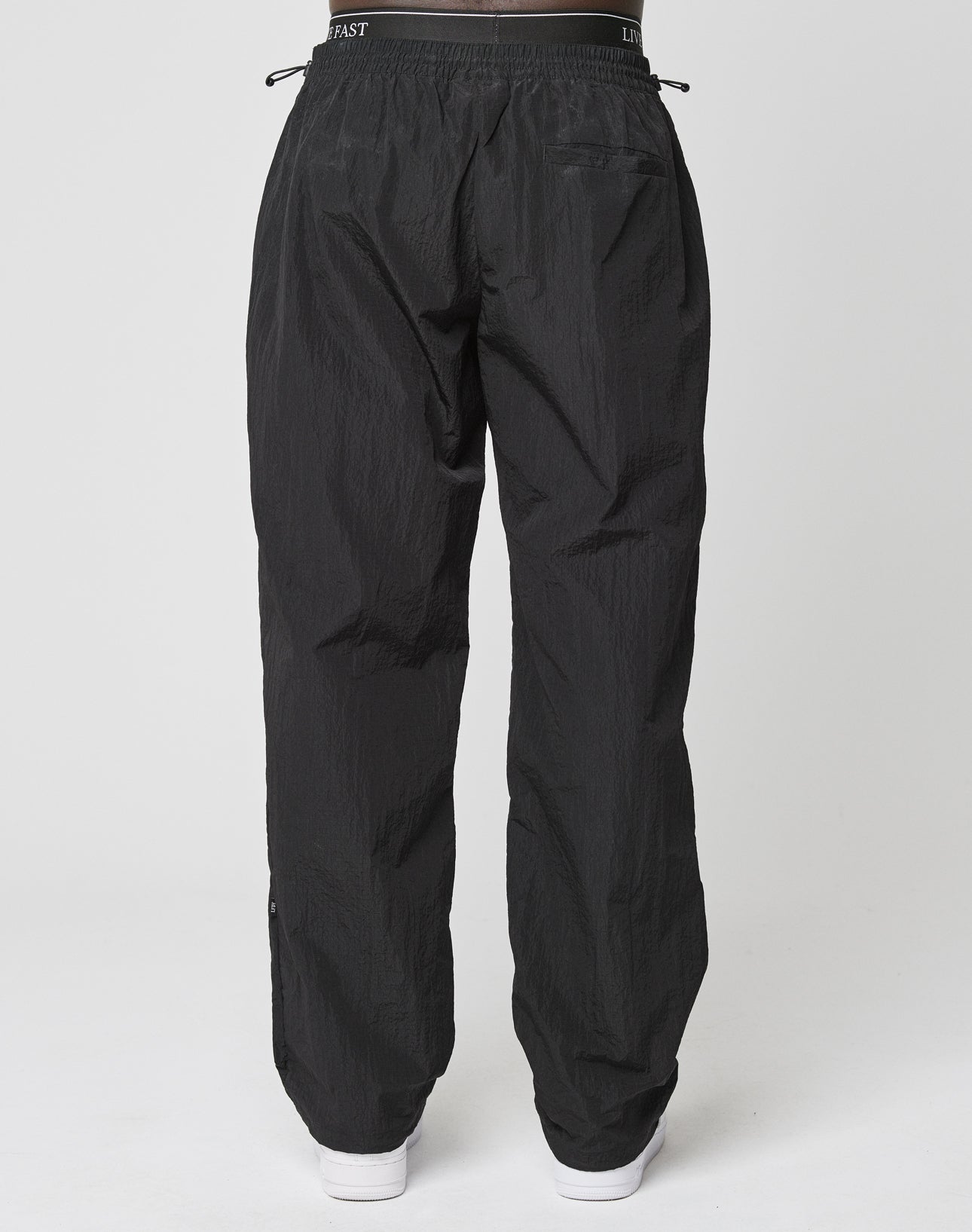 A person is shown from the back wearing the LIVE FAST DIE YOUNG Live Fast Nylon Jogger—loose black track pants with an oversized fit, black elastic waistband, and subtle texture—paired with white shoes against a white background.