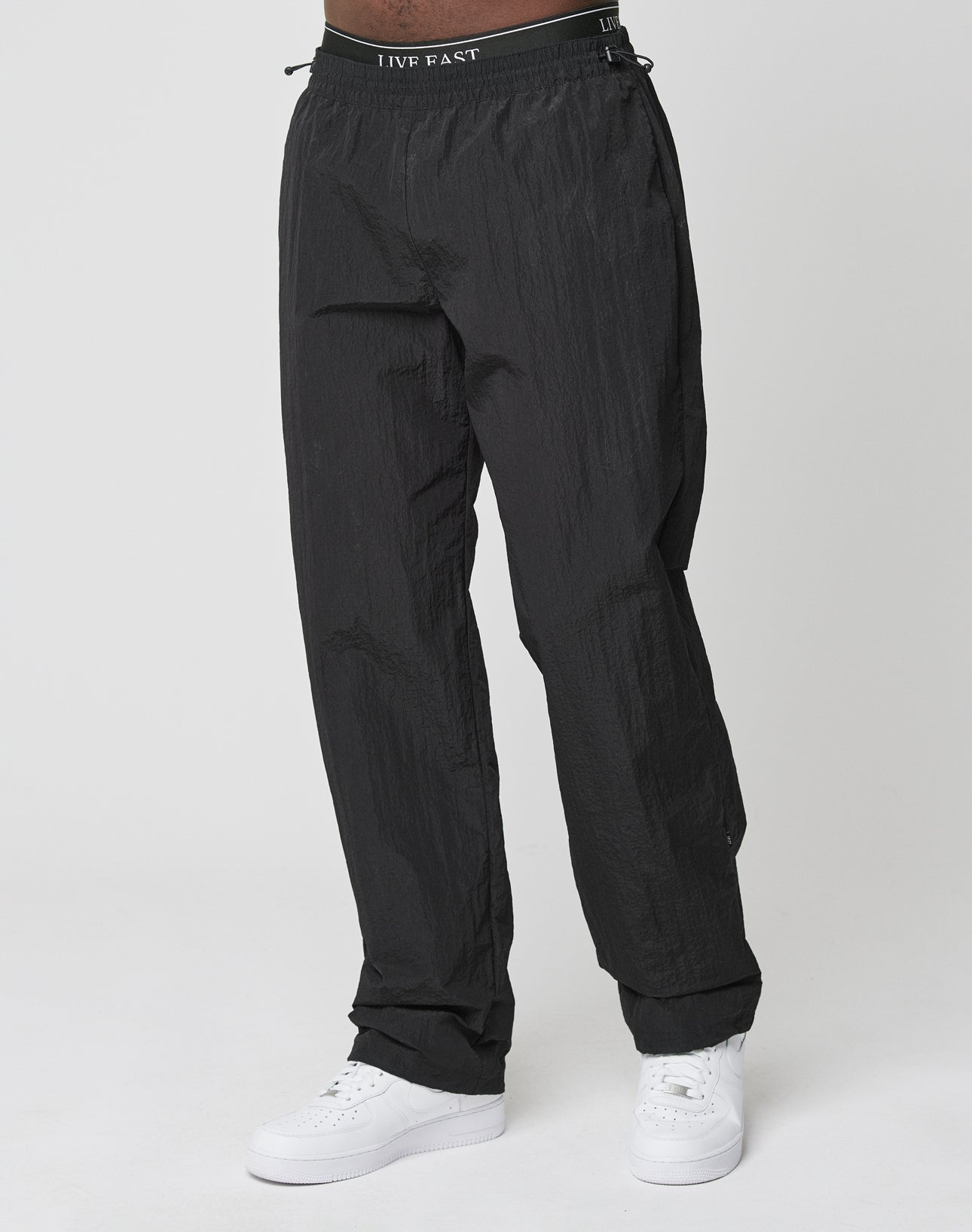 A person wears the LIVE FAST DIE YOUNG Live Fast Nylon Jogger—oversized, loose-fitting black pants with a subtle textured finish and an elastic waistband featuring LIVE FAST text—paired with white sneakers against a plain white background.