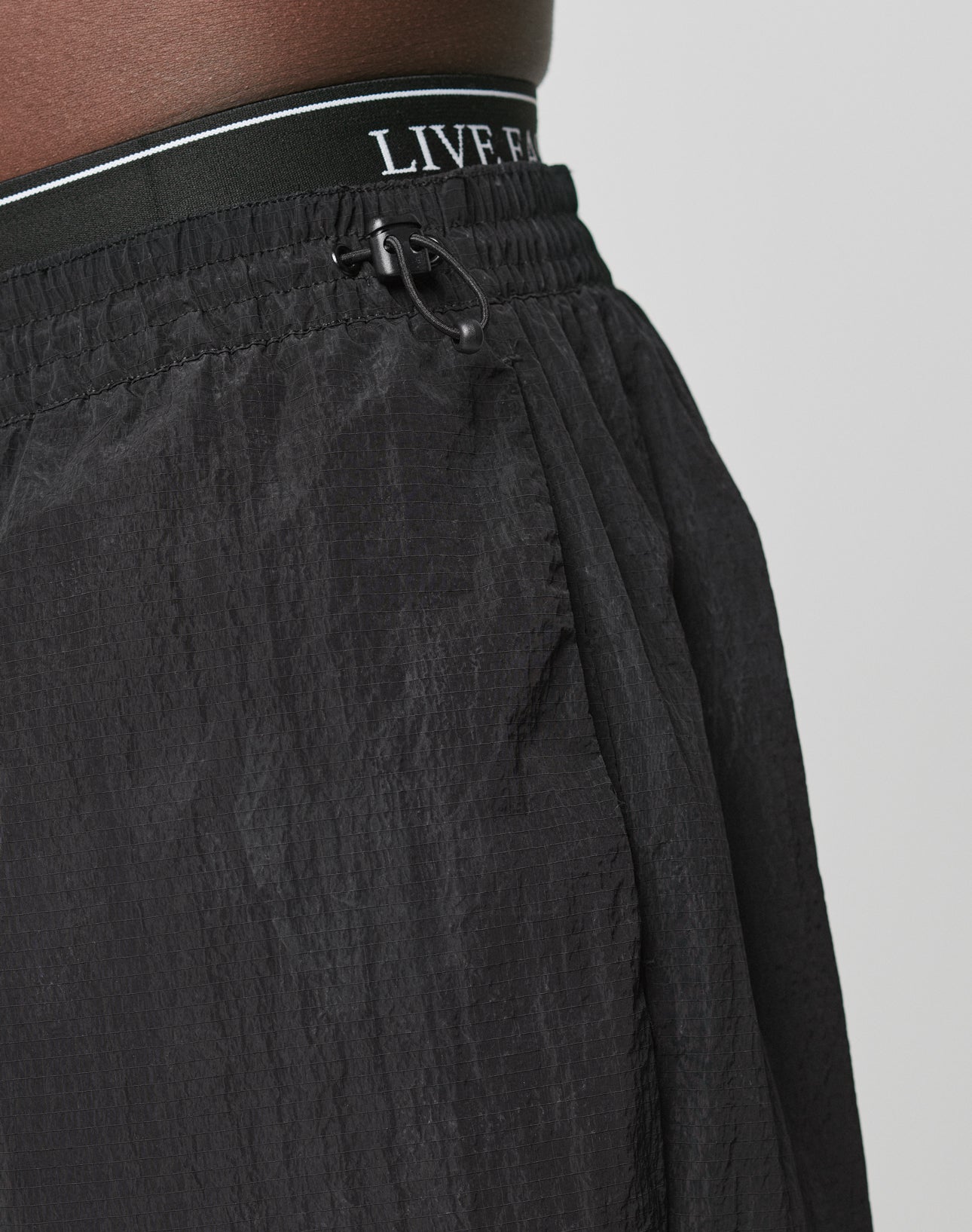 Close-up of a person wearing LIVE FAST DIE YOUNG Live Fast Nylon Jogger, featuring black textured fabric, oversized fit, adjustable drawstring, and an elastikbund waistband with partially visible LIVE F text. Plain light-colored background.