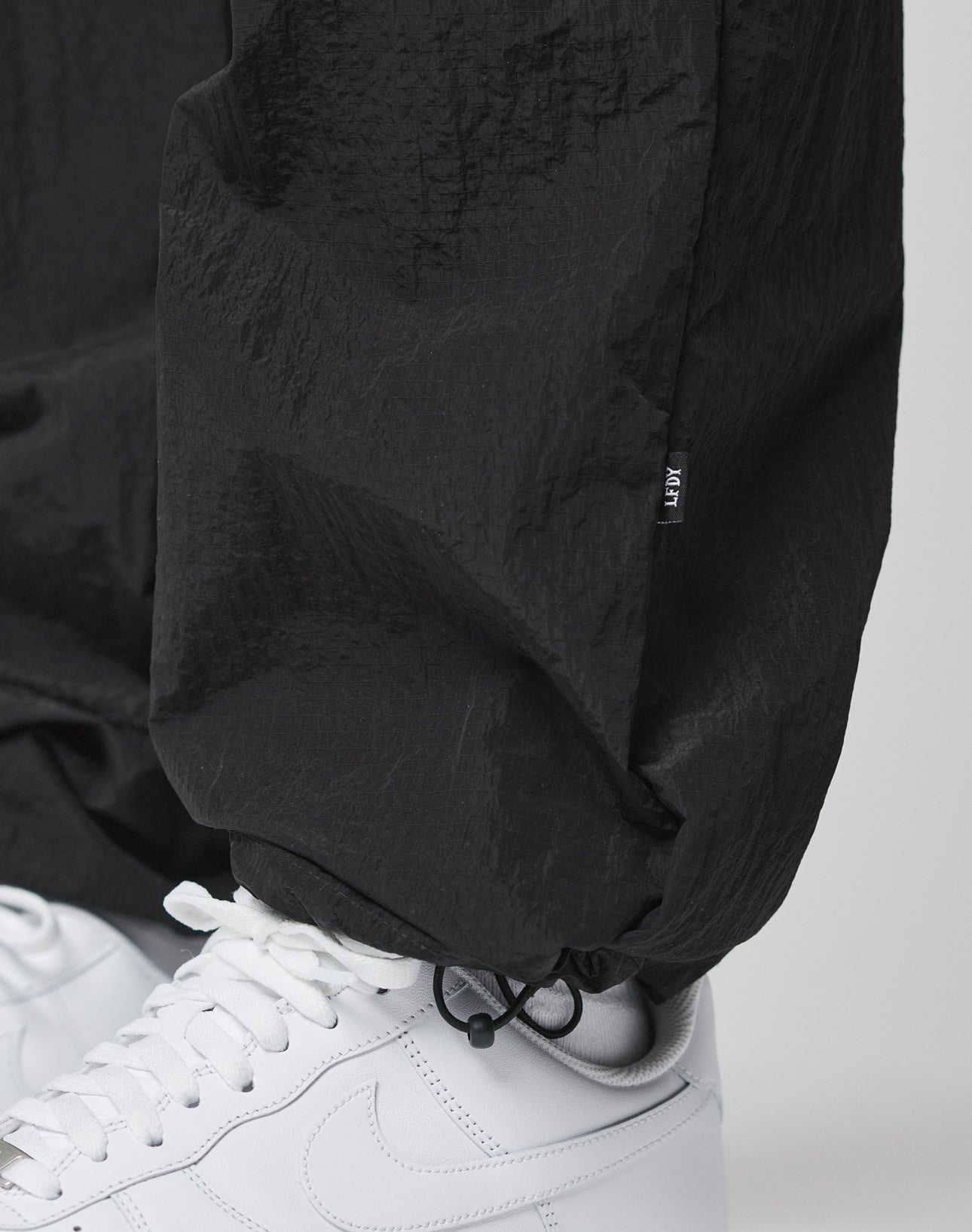 Close-up of a person wearing LIVE FAST DIE YOUNGs Live Fast Nylon Jogger in black, featuring an oversized fit, elastic waistband, adjustable ankle drawstring, and paired with white Nike sneakers. Made from high-quality polyamide for comfort and style.