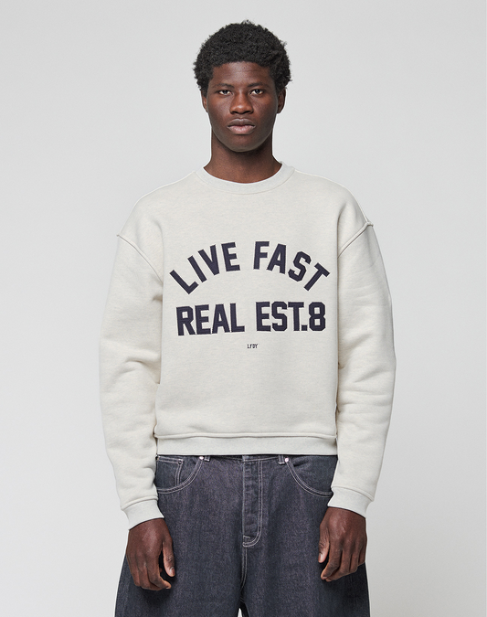 A person stands against a plain background wearing the LFDY Heavyweight Lounge Sweater by LIVE FAST DIE YOUNG, featuring an oversized 100% Baumwolle fit and bold "LIVE FAST REAL EST.8" text on the front, paired with loose dark jeans.