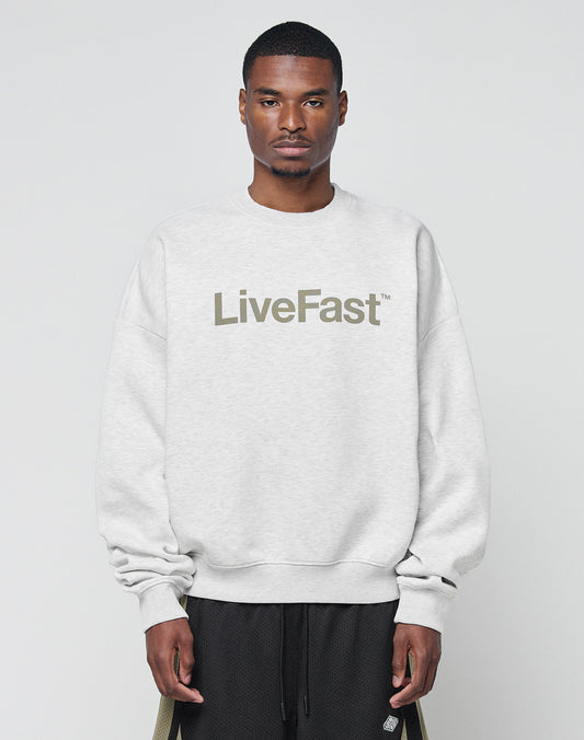 A man wearing the Basic LiveFast Sweater by LIVE FAST DIE YOUNG, a light gray oversize fit made from soft cotton with LiveFast⢠on the front, stands against a plain background, looking straight at the camera with a neutral expression.