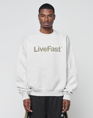 A man wearing the Basic LiveFast Sweater by LIVE FAST DIE YOUNG, a light gray oversize fit made from soft cotton with LiveFast™ on the front, stands against a plain background, looking straight at the camera with a neutral expression.