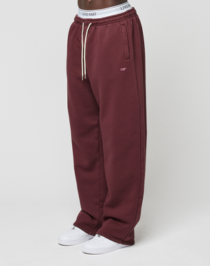 A person wears LIVE FAST DIE YOUNG Basic Wide Jogger in maroon with side pockets, white drawstrings, and a logo-print waistband, paired with white sneakers; only the lower body is visible against a plain background.