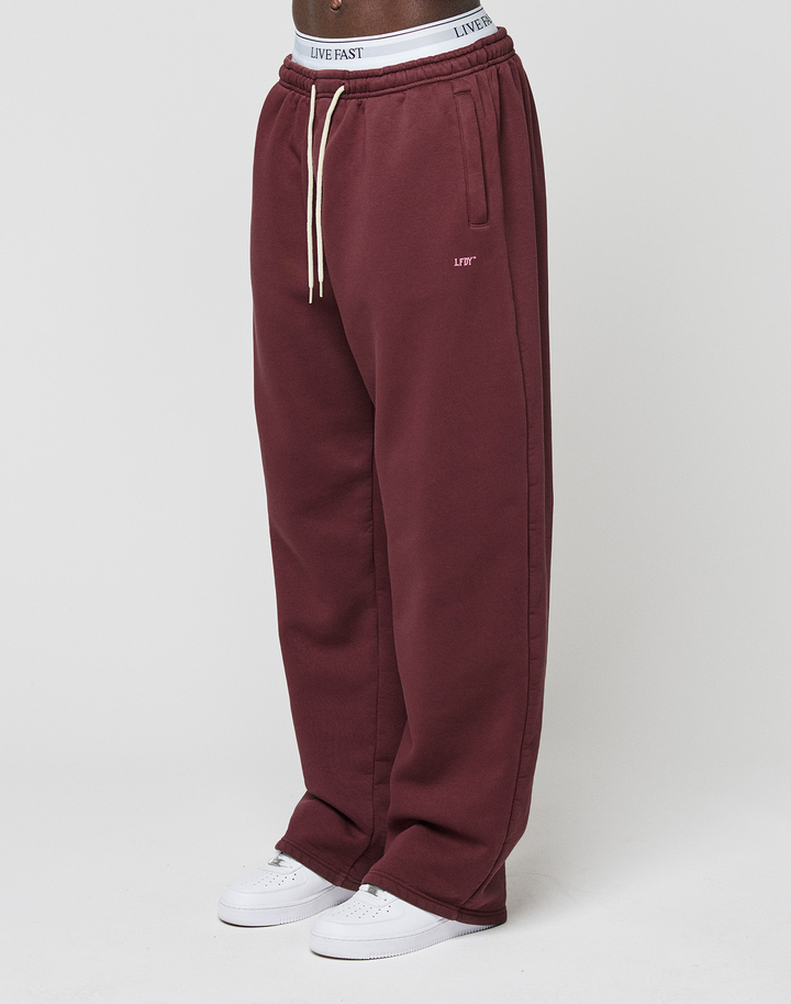 A person wears LIVE FAST DIE YOUNG Basic Wide Jogger in maroon with side pockets, white drawstrings, and a logo-print waistband, paired with white sneakers; only the lower body is visible against a plain background.