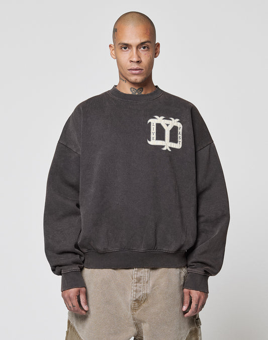 A person with a shaved head wears the Live Fast DY Sweater by LIVE FAST DIE YOUNG, featuring a palm tree and letter P appliqué, paired with light brown jeans against a plain light background.