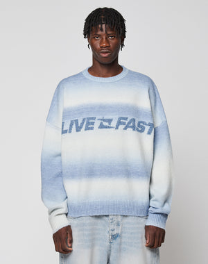 A person wears the Live Fast Boucle Sweater by LIVE FAST DIE YOUNG—an oversized blue and white gradient knit with "LIVE FAST" on the front—paired with light blue jeans, standing against a plain light background.