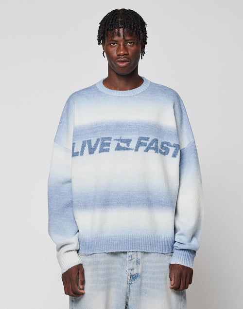 A person wears the Live Fast Boucle Sweater by LIVE FAST DIE YOUNG—an oversized blue and white gradient knit with "LIVE FAST" on the front—paired with light blue jeans, standing against a plain light background.