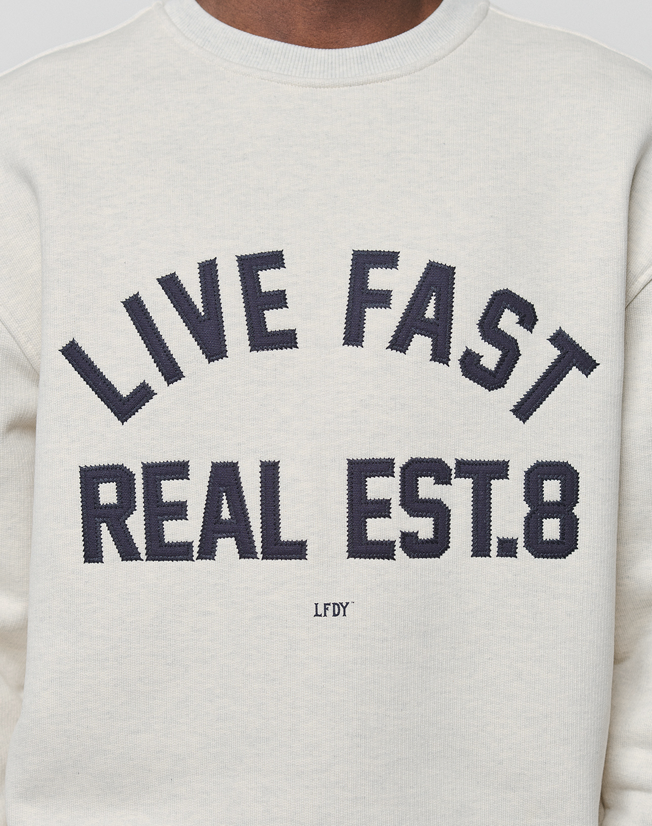 A close-up of the LFDY Heavyweight Lounge Sweater by LIVE FAST DIE YOUNG, made from 100% cotton in a light hue, with bold "LIVE FAST REAL EST.8" lettering and smaller "LFDY" text underneath.