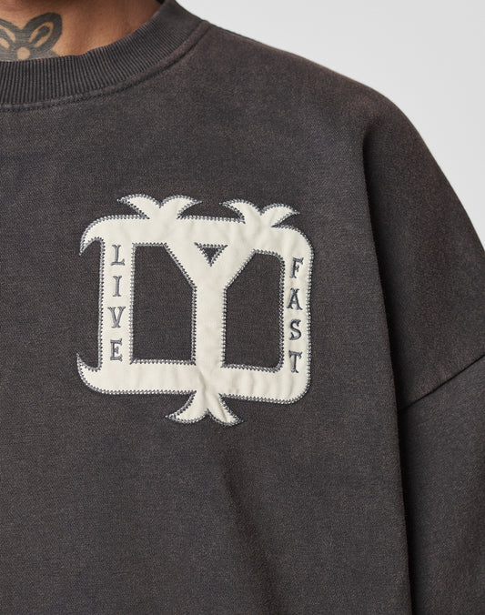 A close-up of the Live Fast DY Sweater by LIVE FAST DIE YOUNG, a dark oversized sweater with white palm tree appliqué and "LIVE FAST" text on the chest. Part of a tattooed neck is visible.
