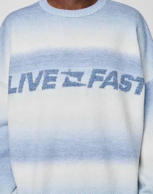 A person wears the LIVE FAST DIE YOUNG Live Fast Boucle Sweater—an oversized fuzzy blue and white gradient knit with angestrickte Bündchen, “LIVE FAST,” and a small plane graphic on the chest.