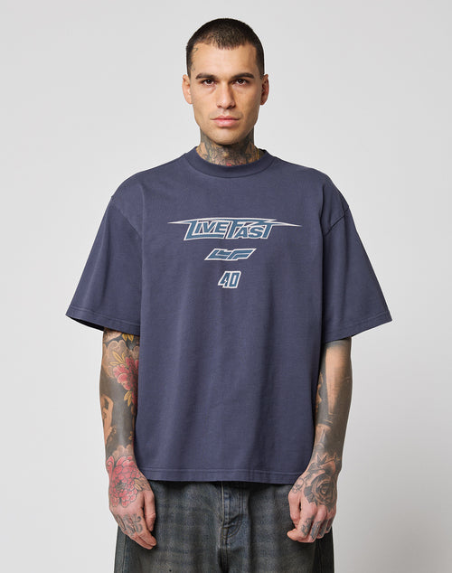 A person with short hair and visible tattoos wears an oversize fit, dark navy LFDY Triple Racing Tee by LIVE FAST DIE YOUNG with a Multilogo Frontprint reading LIVE FAST CF 40, standing against a plain light gray background.