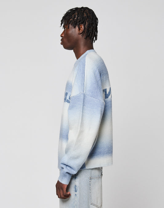 A person with short, textured hair stands in profile, wearing the Live Fast Boucle Sweater from LIVE FAST DIE YOUNG—a blue and white gradient, oversize knit with angestrickte Bündchen—paired with light-wash jeans, against a plain background.