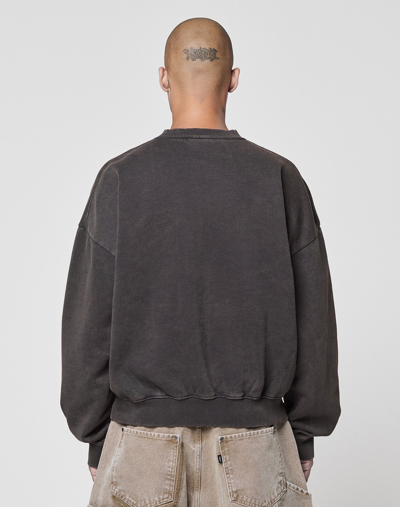 A person with a shaved head and a neck tattoo is shown from behind, wearing the faded black Live Fast DY Sweater by LIVE FAST DIE YOUNG and light brown, worn-out jeans against a plain light background.