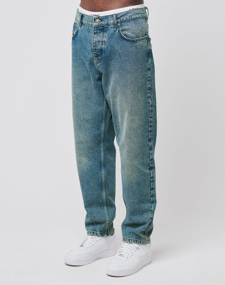 Wearing LIVE FAST DIE YOUNG Regular Denim blue jeans, made from recycled cotton with a regular fit and tapered leg, paired with white sneakers. Only the lower torso and legs appear against a plain white background. | model