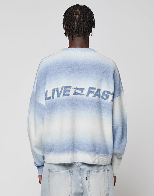 A person with short, twisted hair faces away, wearing the LIVE FAST DIE YOUNG Live Fast Boucle Sweater in a blue and white gradient with "LIVE FAST" on the back, paired with light blue jeans with Angestrickte Bündchen.