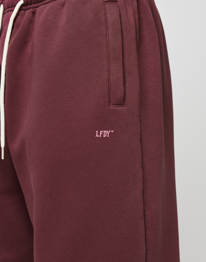 Close-up of a person wearing the LIVE FAST DIE YOUNG Basic Wide Jogger in maroon with wide fit, side pockets, white drawstrings, and small white LFDY™ logo embroidered on the left thigh.