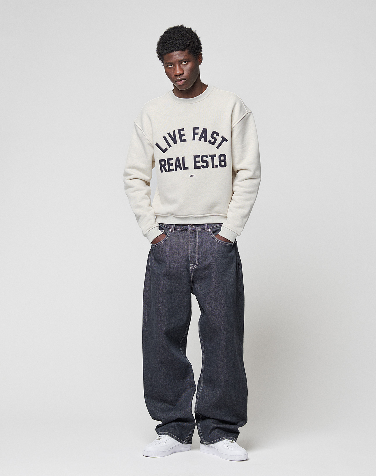 A person stands against a plain light background, wearing the LIVE FAST DIE YOUNG LFDY Heavyweight Lounge Sweater with bold lettering, paired with loose dark jeans and white sneakers. Hands in pockets, they look at the camera.
